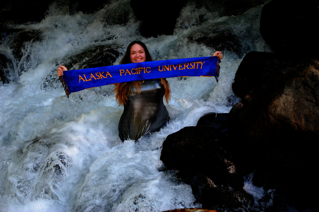 Miriam Mezzetti holding an Alaska Pacific University banner in a rushing river.