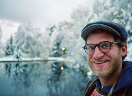 Smiling bearded man in flat cap and glasses - frost-covered trees and pond in background.