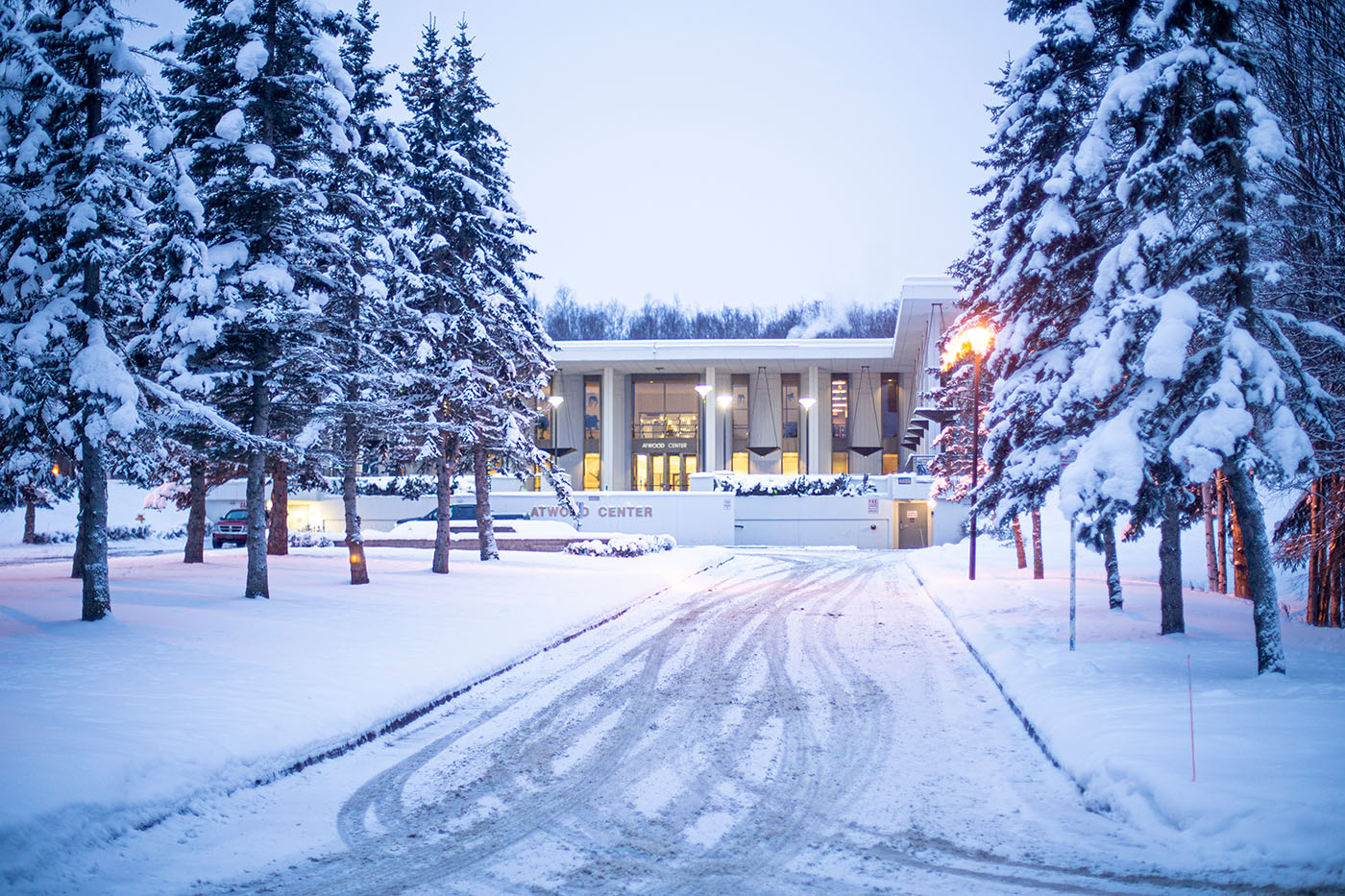 Snow-covered pathway leading to the Atwood Center building, flanked by snow-laden evergreen trees under a twilight sky.