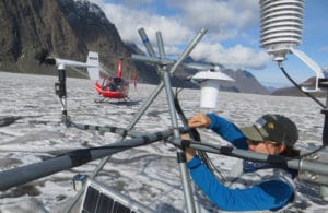 A researcher assembles a monitoring system on the glacier, with a helicopter in the background. Photo credit: Jason Geck