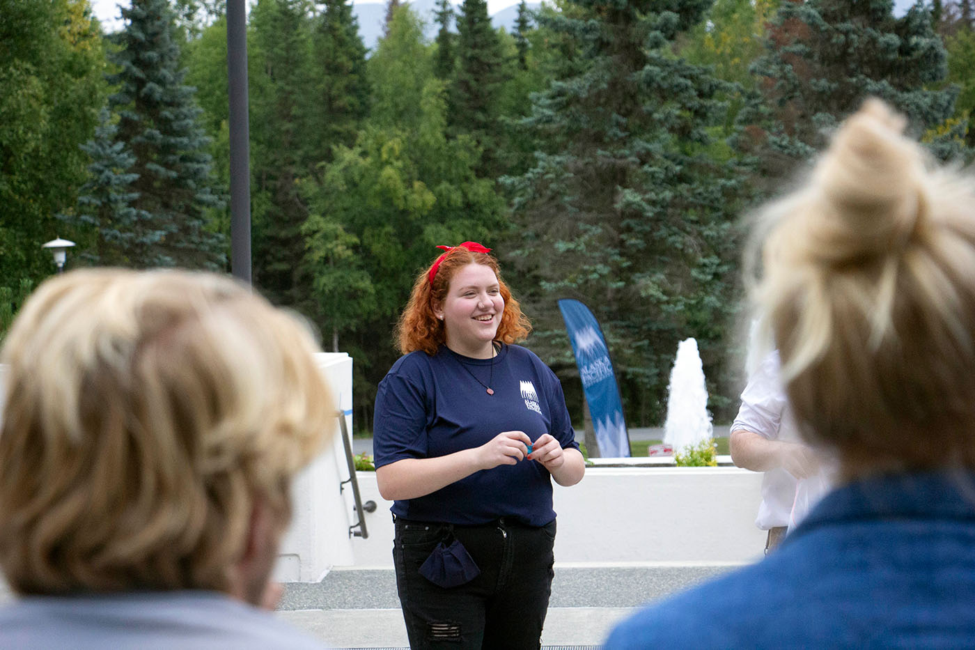 A person with red hair wearing a blue shirt and red headband speaks to an audience, with a fountain and trees in the background.