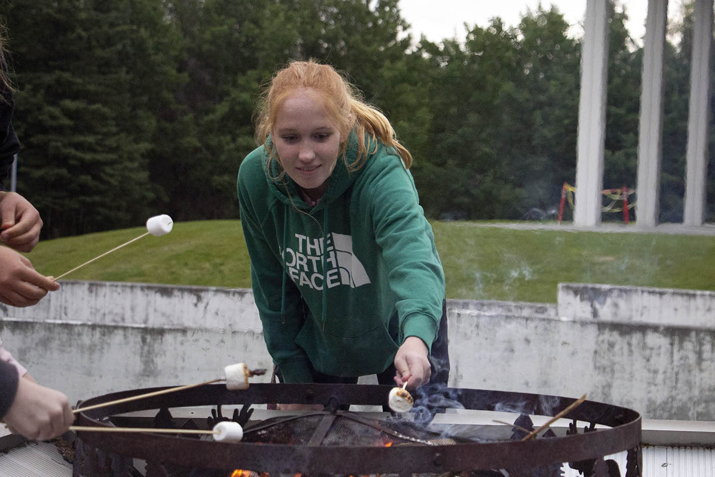 A person roasting marshmallows over a fire pit outdoors, wearing a green hoodie, with a grassy area and tall pillars in the background.