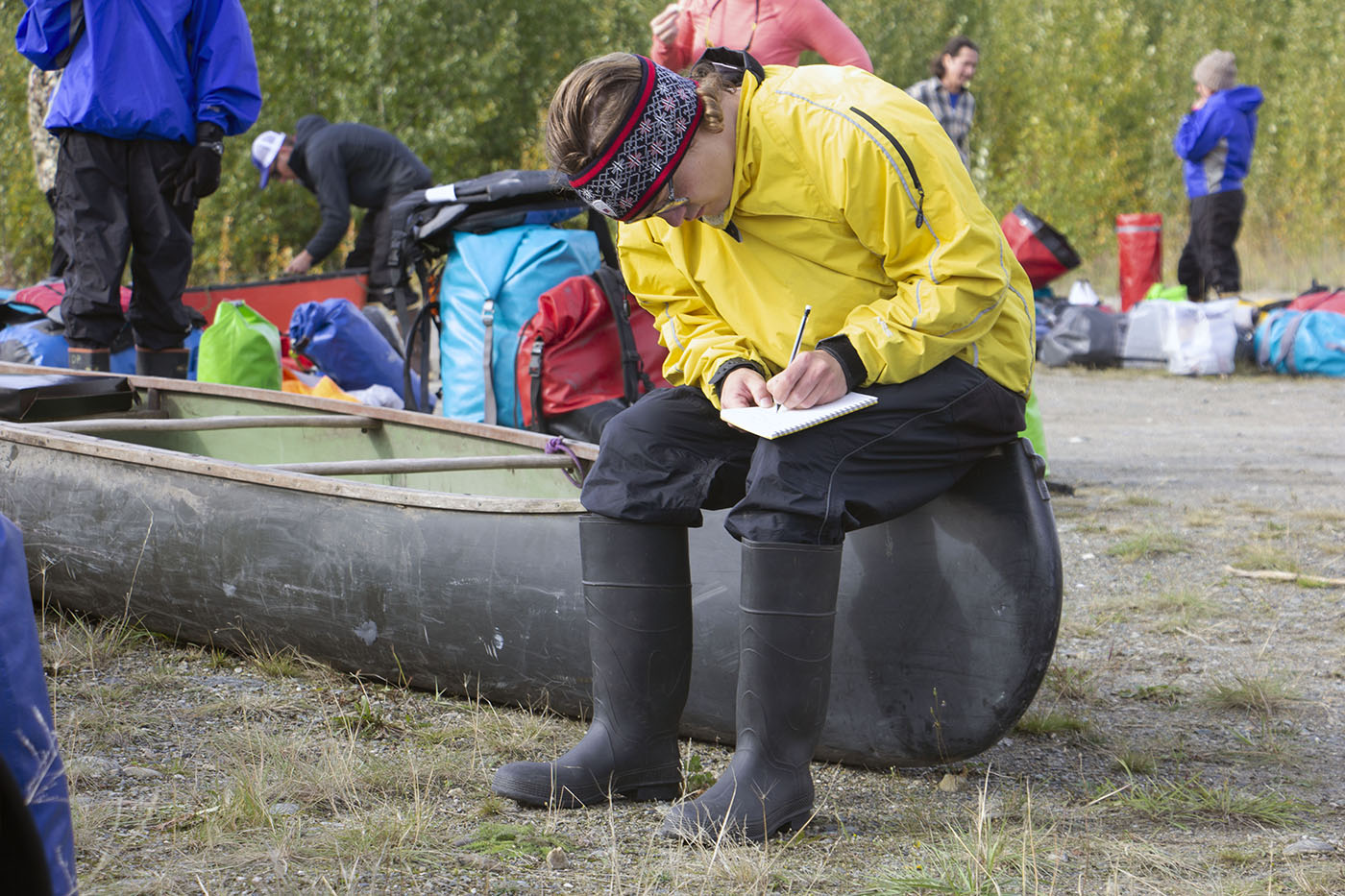 A person in a yellow raincoat and black boots sitting on a canoe, writing in a notebook in an outdoor setting with other people and gear in the background.