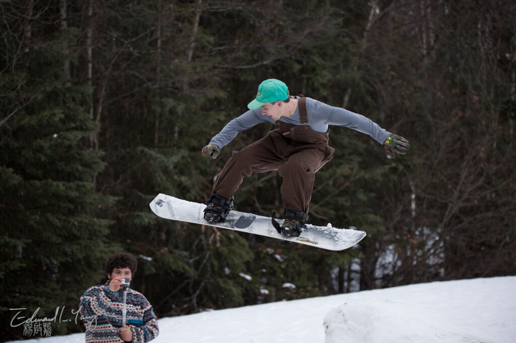 A person performs a jump on a snowboard with a forested snowy hill in the background, another person watches.