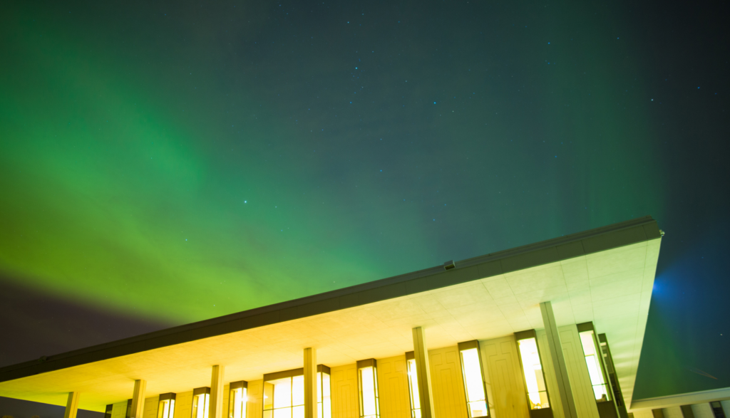 A modern building with large windows, illuminated at night with a green aurora borealis glowing in the sky above.