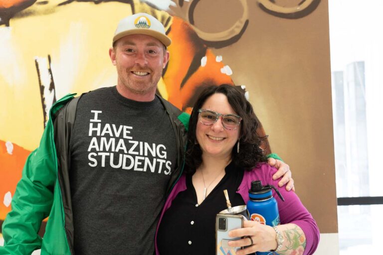 Two people standing together and smiling, with one wearing a shirt that says "I HAVE AMAZING STUDENTS" and a green jacket, and the other holding a water bottle and a phone. They are in front of a colorful mural.
