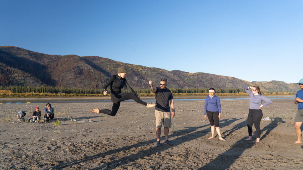 A person mid-air performing a high kick, with others standing and watching on a sandy riverbank, mountains in the background under a clear blue sky.