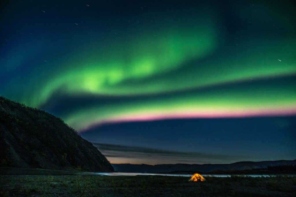 A tent illuminated under a vibrant aurora borealis display with green and pink hues in the night sky, set against a backdrop of mountains and a body of water.
