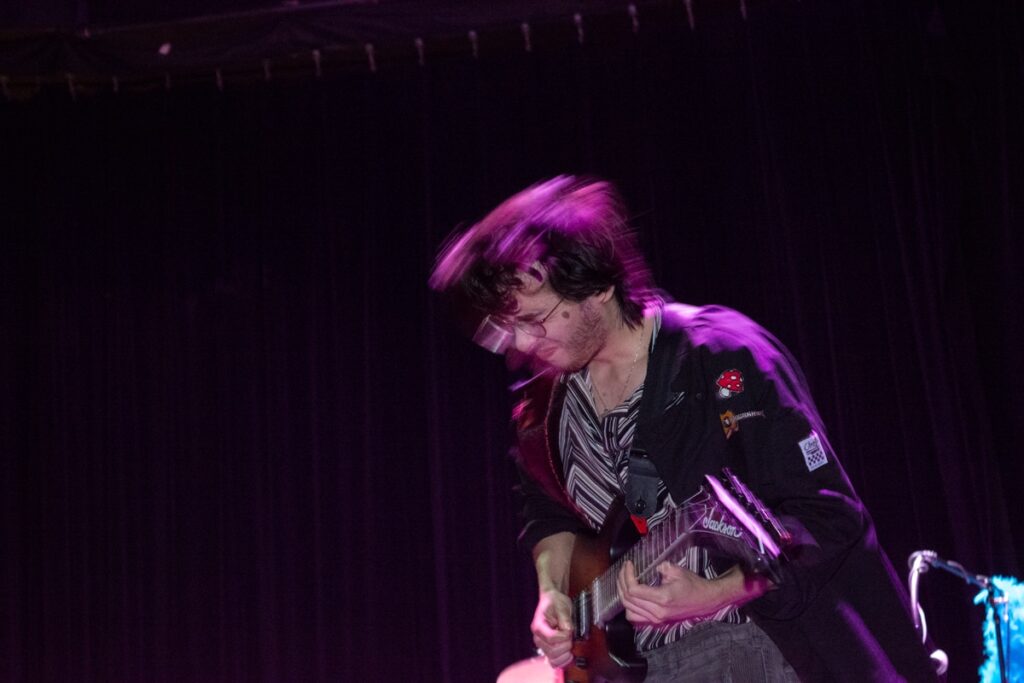 A lead guitarist with long hair plays a Jackson guitar on stage, wearing a black jacket with patches, while other band members perform with a Korg keyboard, drums, and bass guitar under purple stage lighting.