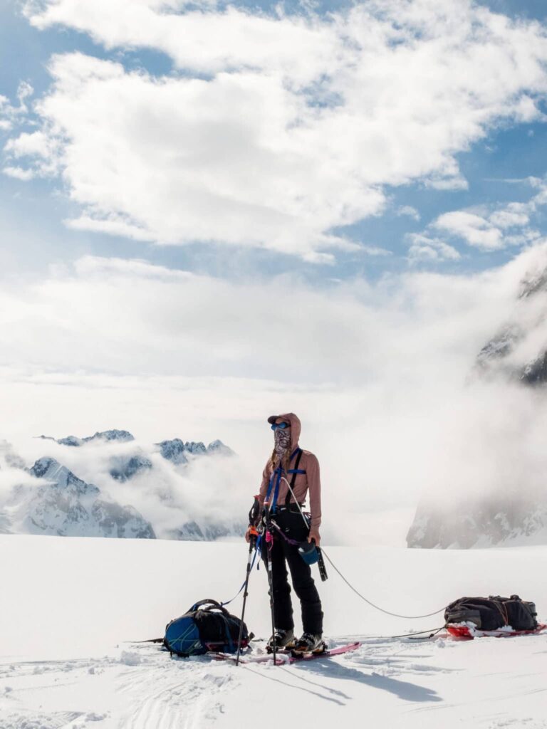 A person on skis with climbing gear and backpacks in a snowy mountainous area with clouds and peaks in the background.