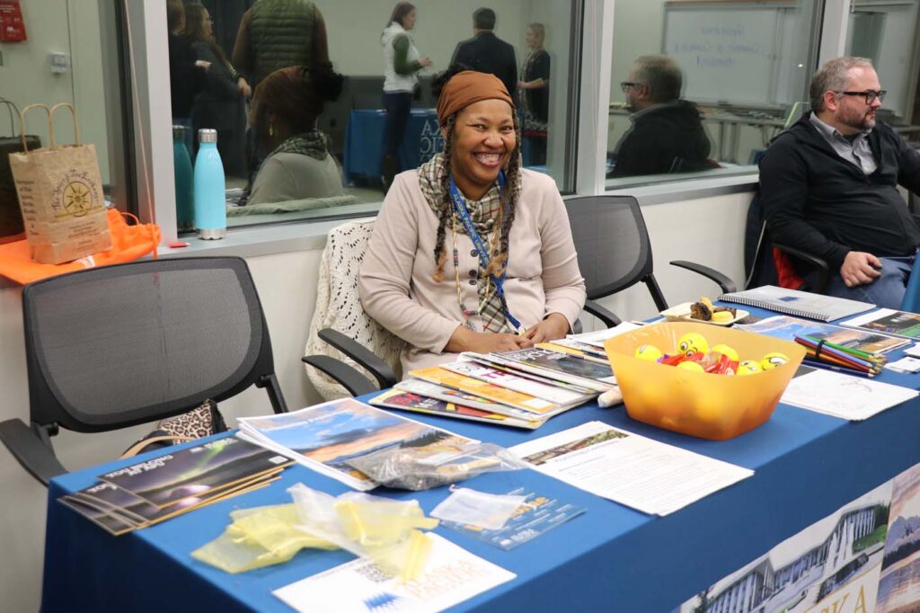 A woman sitting at a blue-draped table with brochures and a bowl of yellow knickknacks, smiling and holding a pen, with a window and other people in the background.