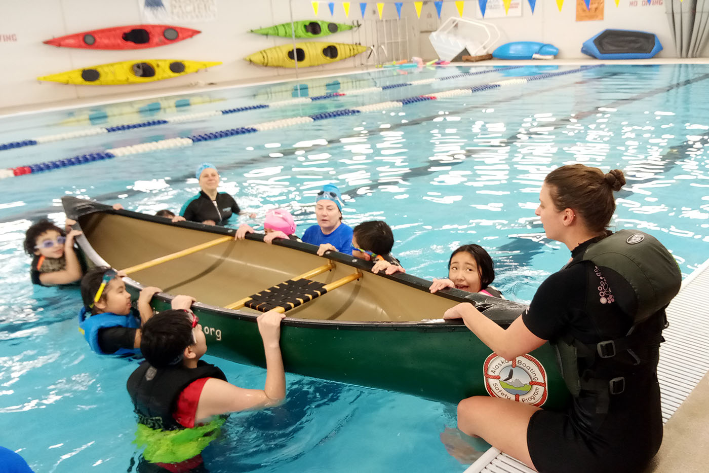 An instructor and several children in life jackets learning to canoe in a pool, with kayaks and colorful flags on the wall.