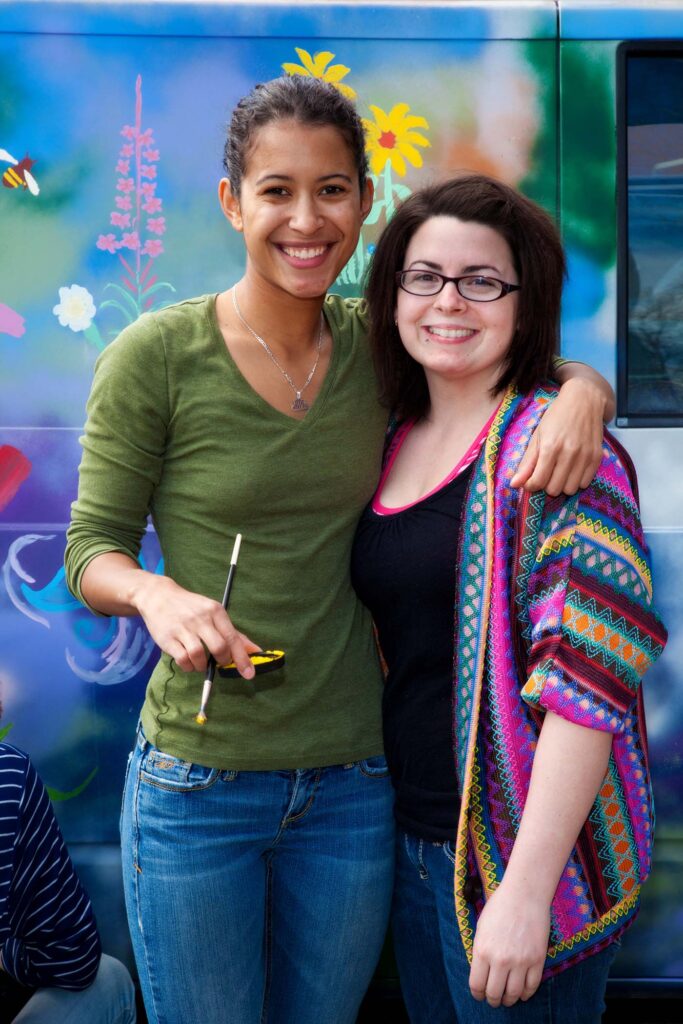 Two women smile and pose for a photo in front of a colorful mural featuring flowers and a bee.