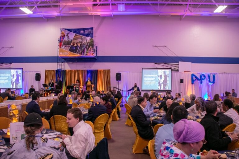 A large group of people dining and listening to a speaker in a decorated indoor venue with APU signage.