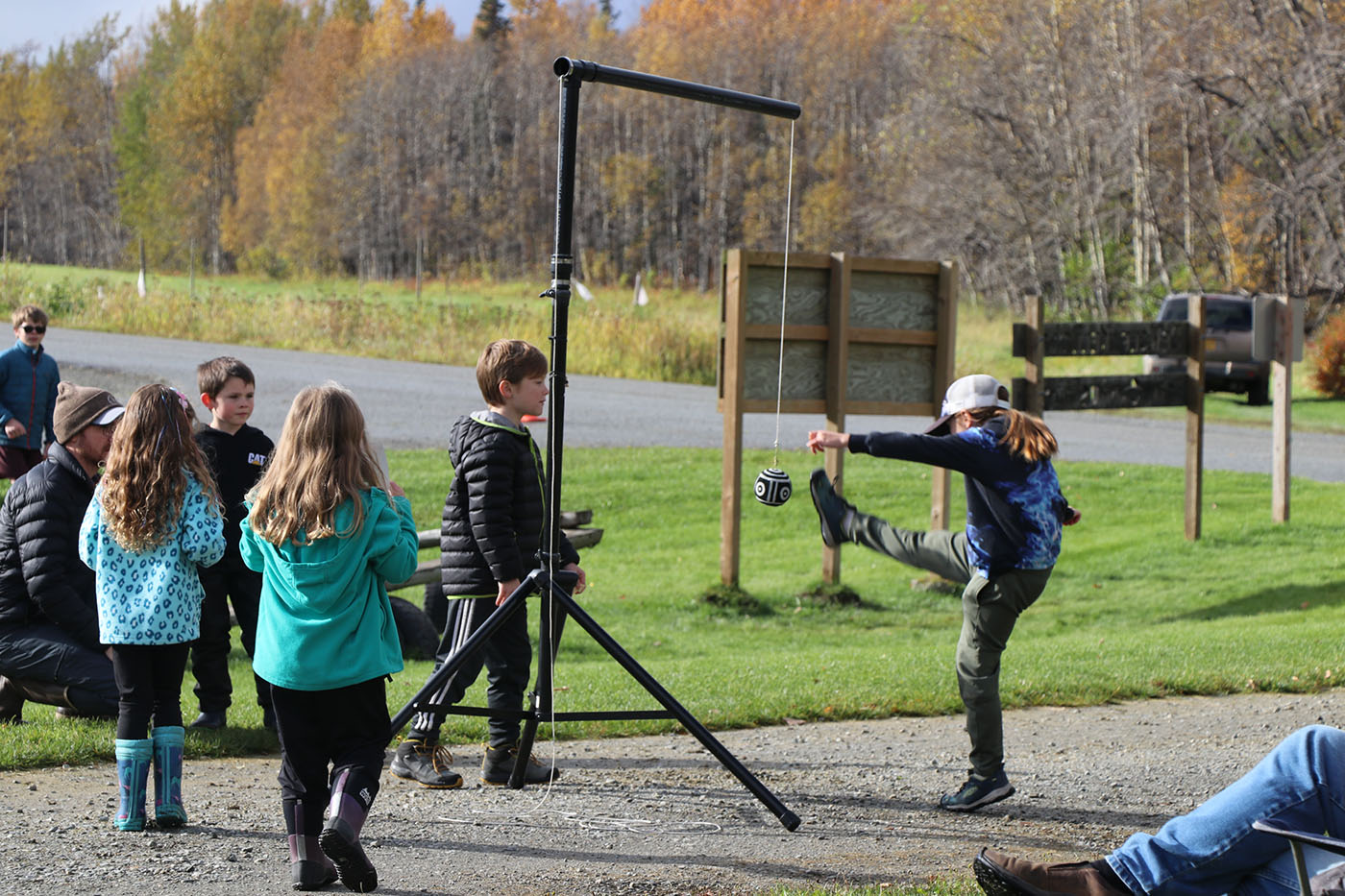 A child kicking a ball in a pendulum game, with other children watching in a grassy area with autumn trees.