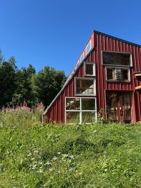 A red modern house with large windows, surrounded by green grass and trees under a clear blue sky.