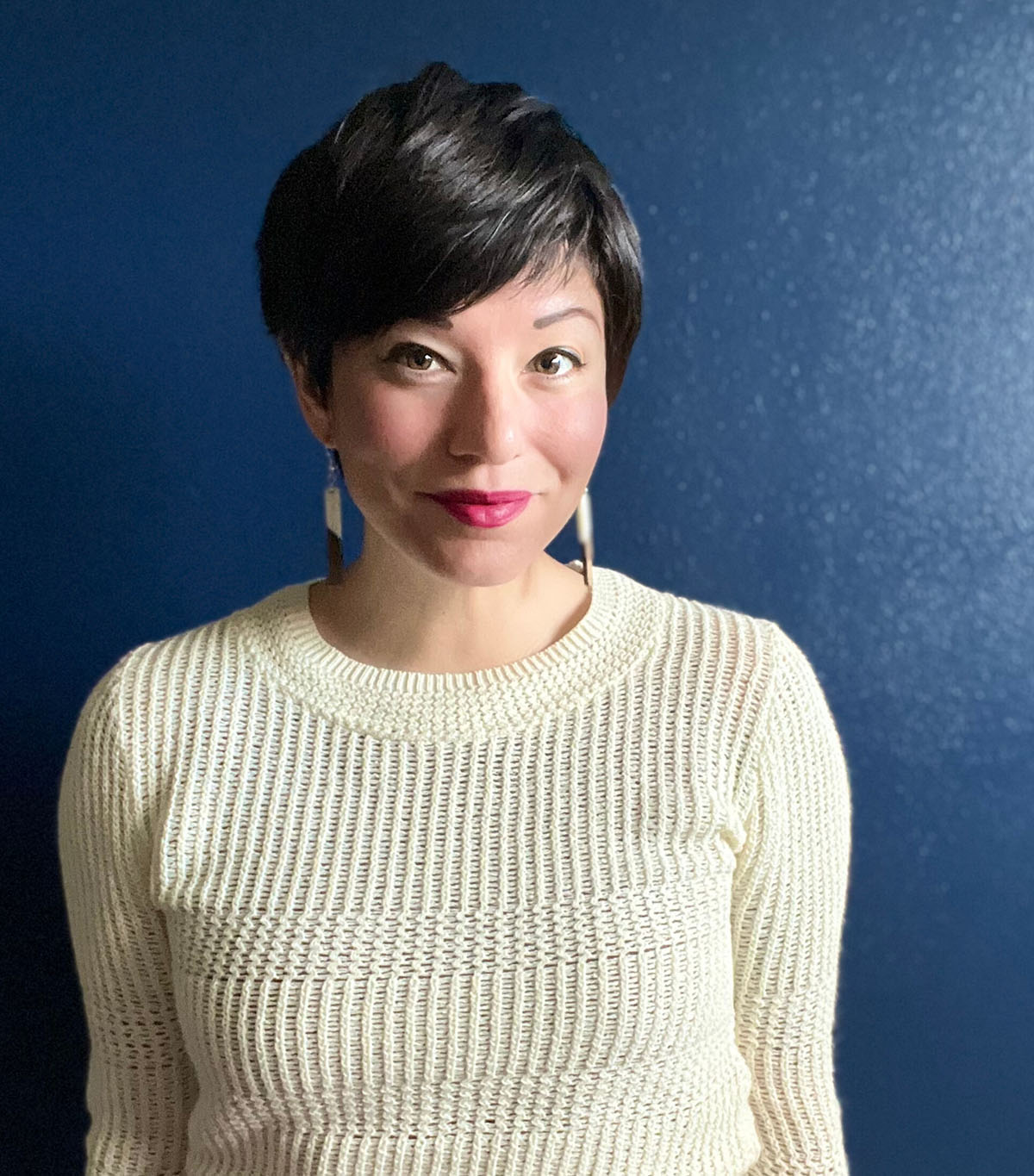 A woman posing indoors against a blue wall, wearing a white knitted sweater and earrings.