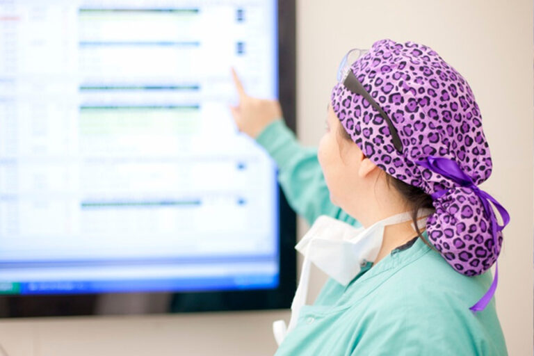 A medical professional in a green outfit with a purple leopard-print cap pointing at a computer screen displaying a list.