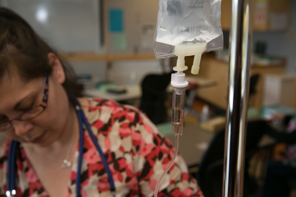 A medical professional in a floral scrub top adjusting an IV drip bag in a clinical setting.