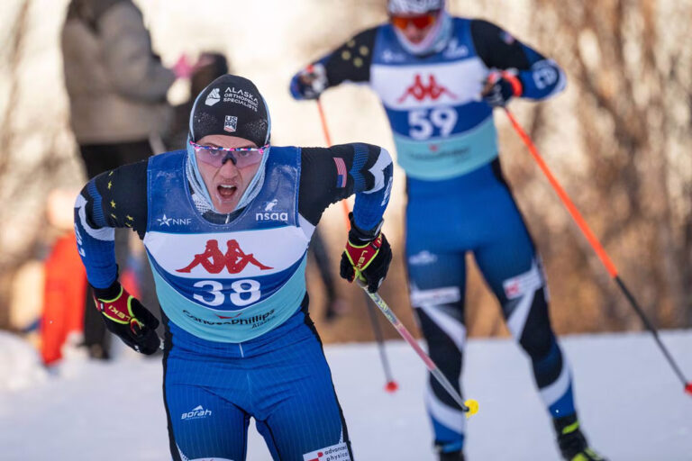 Two cross-country skiers in action, wearing numbered bibs and skiing gear, with a snowy landscape in the background.