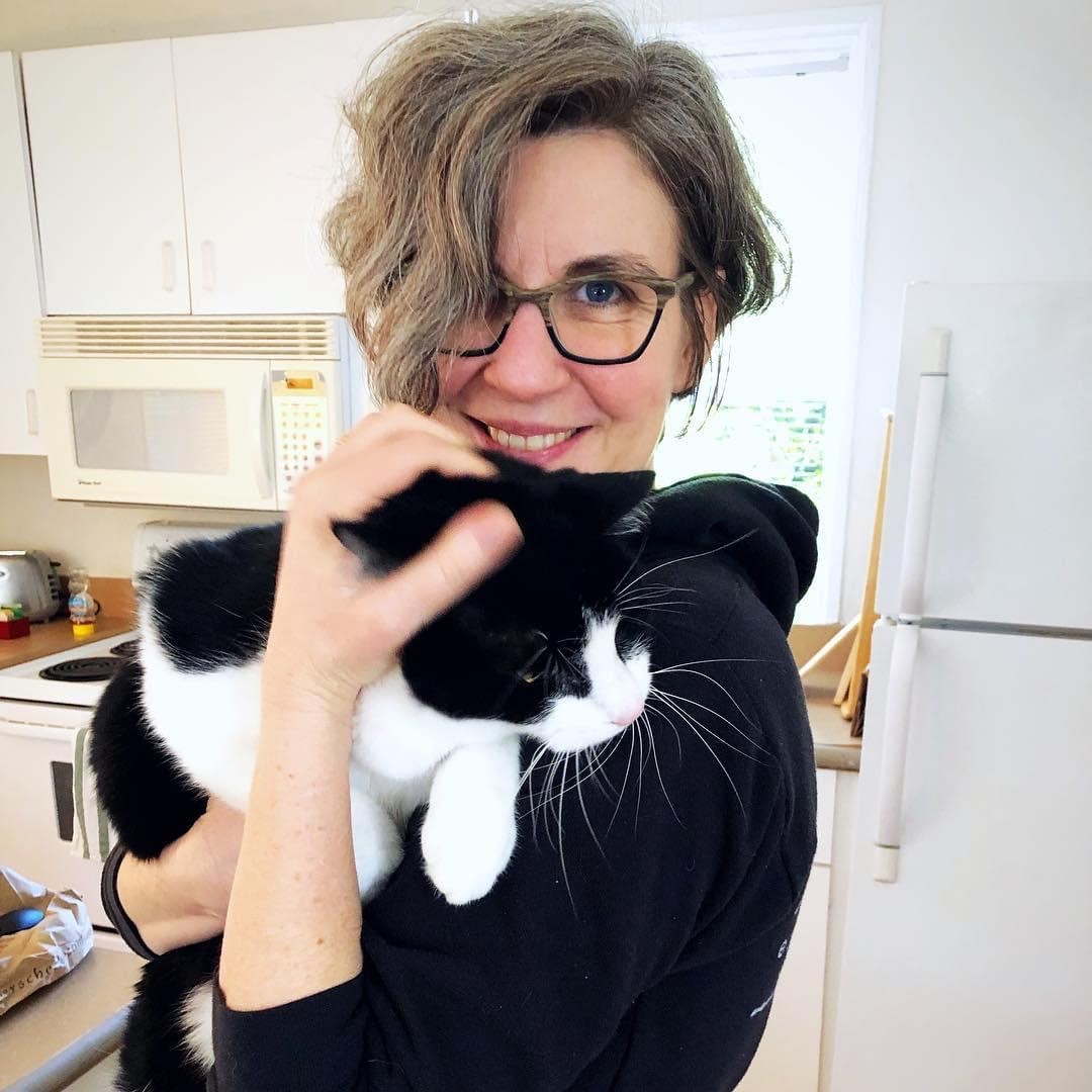 A woman in a black hoodie holding a black-and-white cat in a kitchen setting.