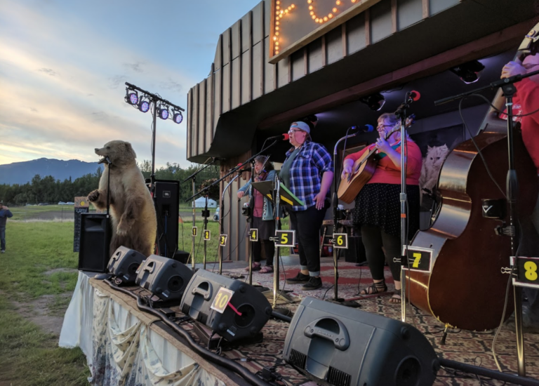 A band performing on a stage with a bear statue and musical instruments, set against a backdrop with mountains.