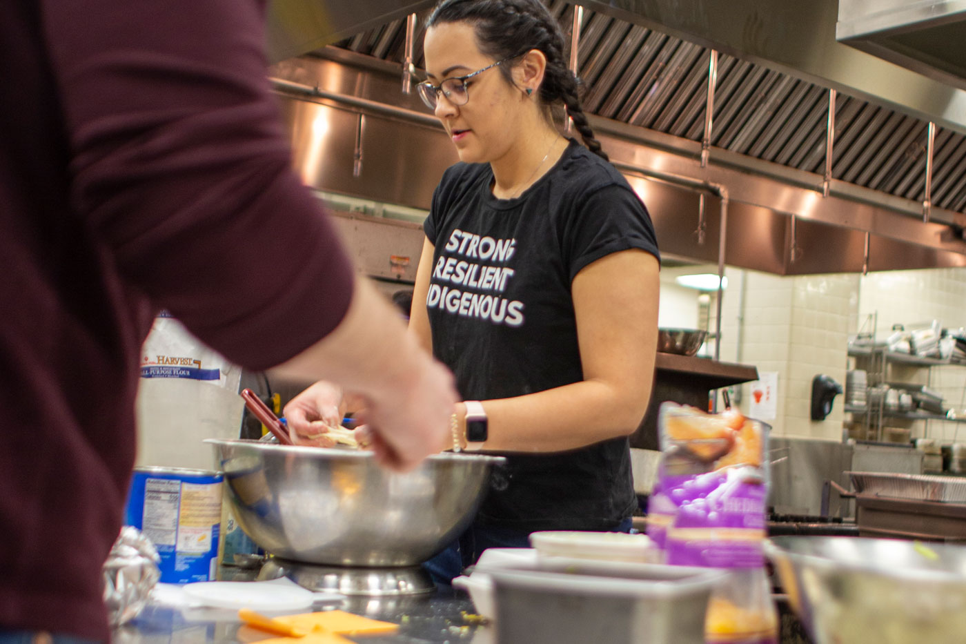 A person in a black shirt with "Strong Resilient Indigenous" text mixing ingredients in a bowl in a professional kitchen.