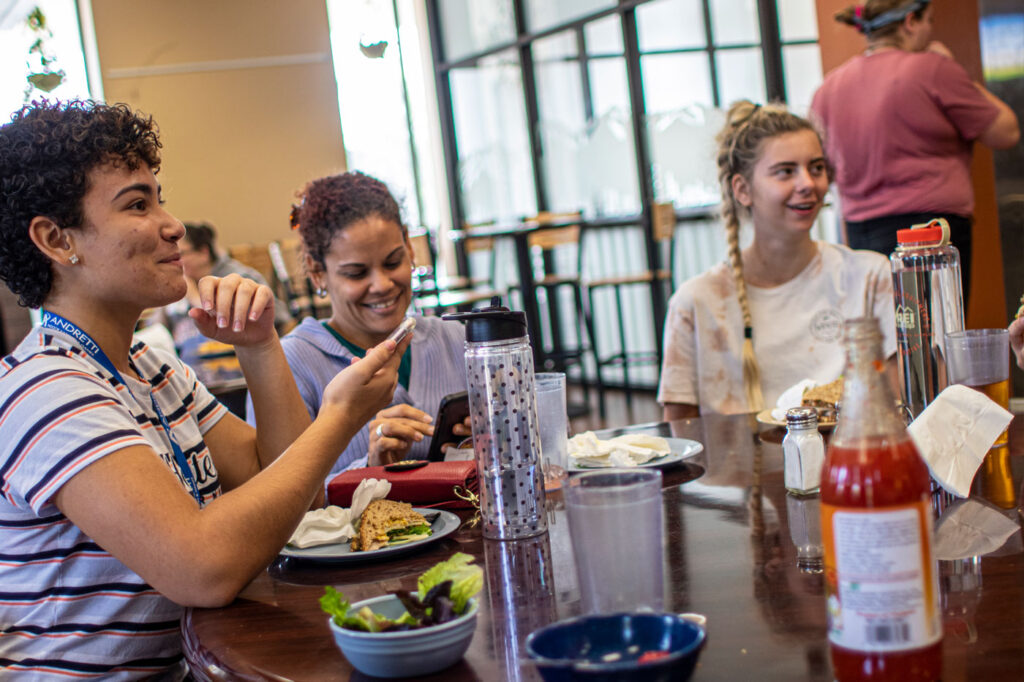 Three young women sitting and smiling at a table, with one using a smartphone, in a casual dining setting.