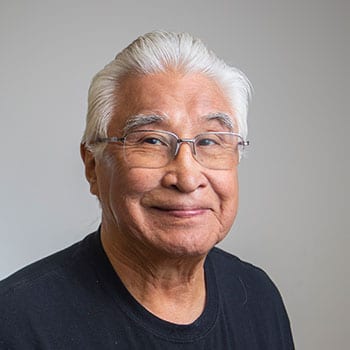An older man with white hair and glasses wearing a black shirt, smiling against a plain background.
