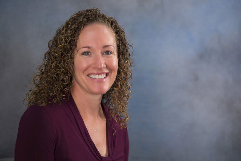 A portrait of a smiling woman with curly hair, wearing a purple top, against a blurred blue background.