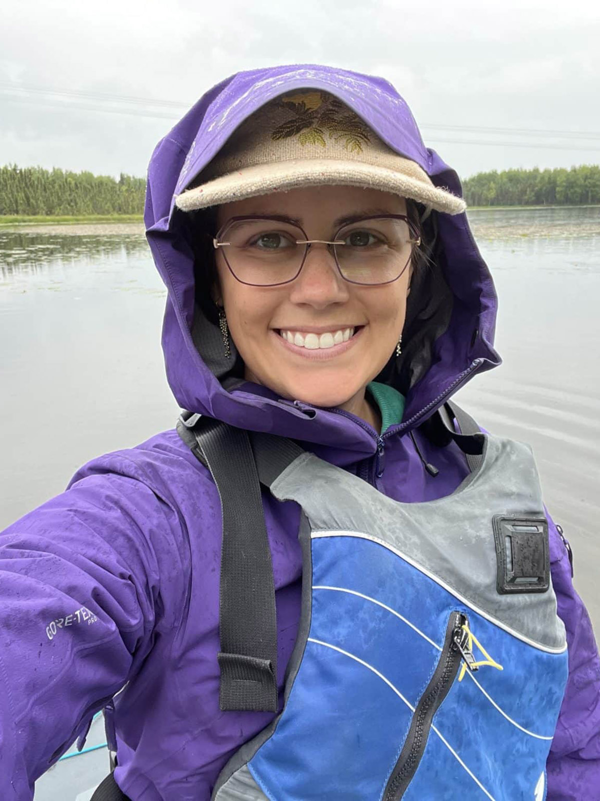 A person in a purple jacket and life vest taking a selfie while kayaking on a lake with trees in the background.
