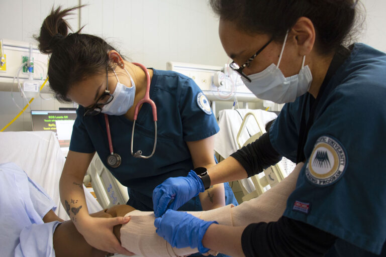 Two medical professionals in blue scrubs and masks tending to a patient, one bandaging an arm, in a hospital setting with medical equipment.