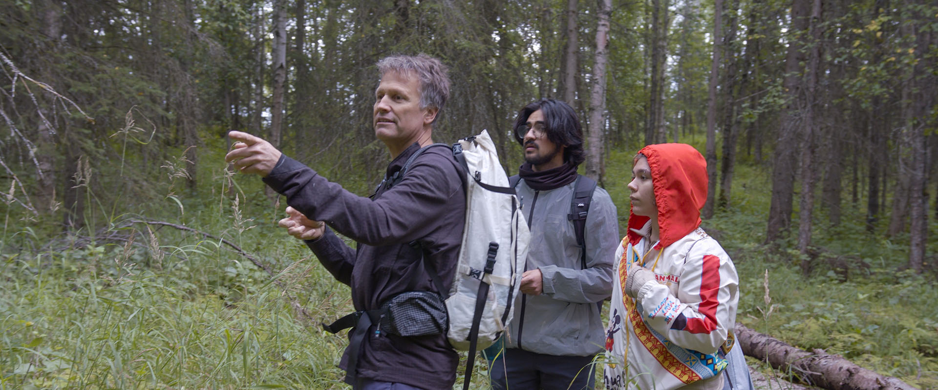 Three people hiking in a forest, with one pointing and the others following, all wearing backpacks.