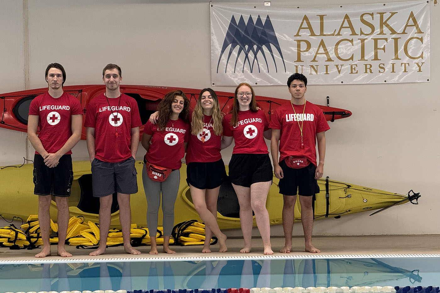 A group of six lifeguards wearing red "LIFEGUARD" shirts with white crosses, standing barefoot by a pool with kayaks and an Alaska Pacific University banner in the background.
